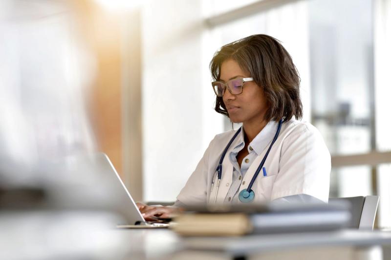 Médecin travaillant sur un ordinateur portable, avec un stéthoscope autour du cou, dans un environnement lumineux de type hôpital ou cabinet médical.