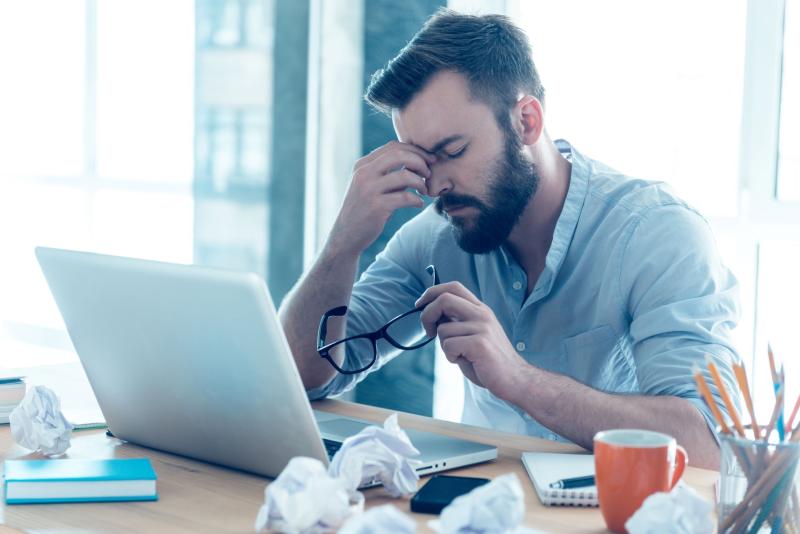 Homme barbu assis à son bureau, visiblement stressé ou fatigué, retirant ses lunettes et se frottant les yeux devant son ordinateur portable, entouré de feuilles froissées.