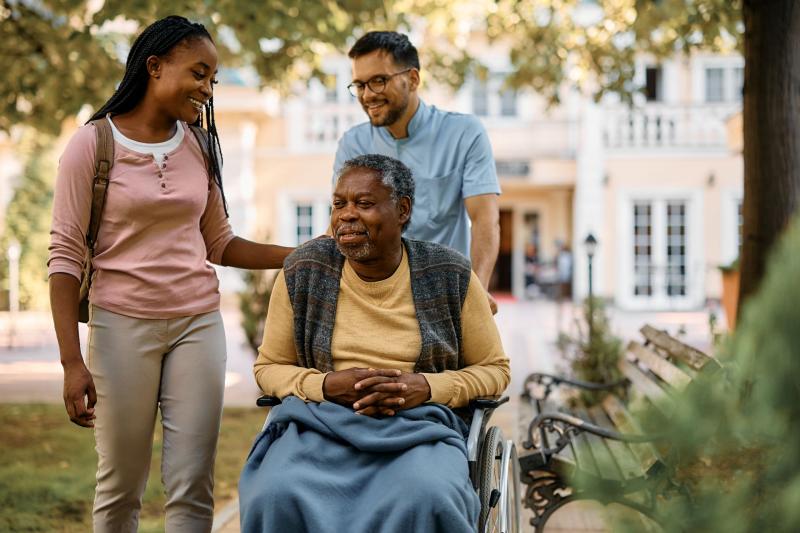 Une personne âgée en fauteuil roulant accompagnée par deux aidants lors d’une promenade en extérieur
