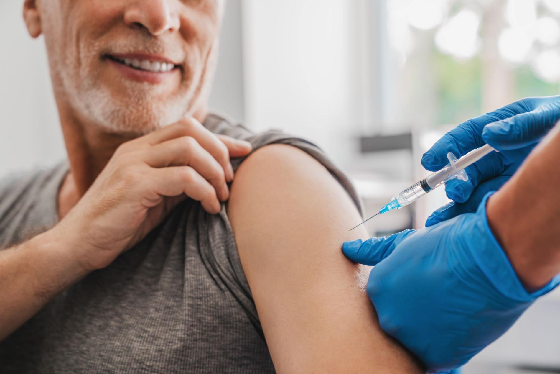 Homme souriant recevant une injection dans le bras par un professionnel de santé portant des gants bleus, illustrant un acte de vaccination