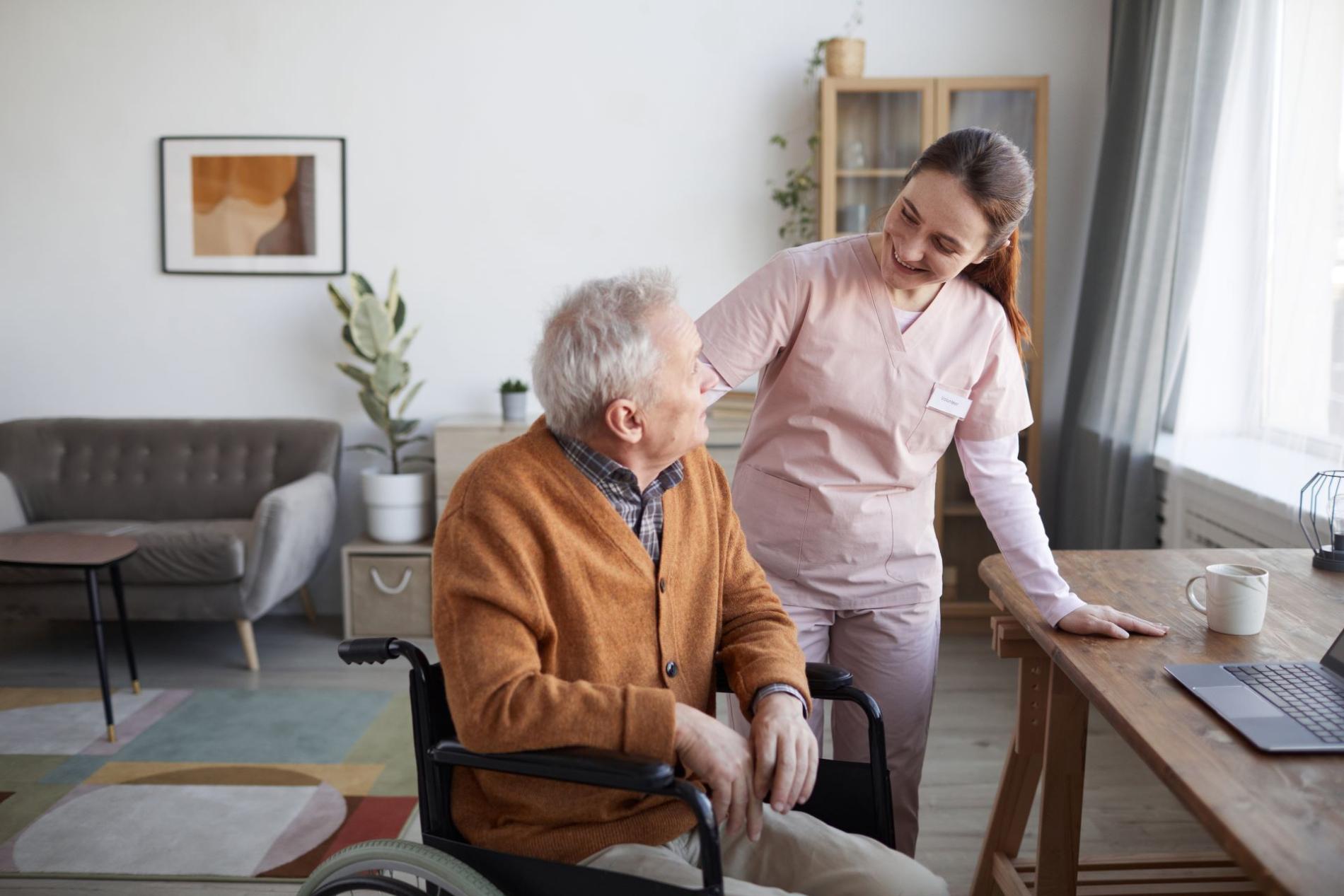 Soignante souriante échangeant avec une personne âgée en fauteuil roulant dans un intérieur chaleureux, illustrant les soins à domicile ou en institution.