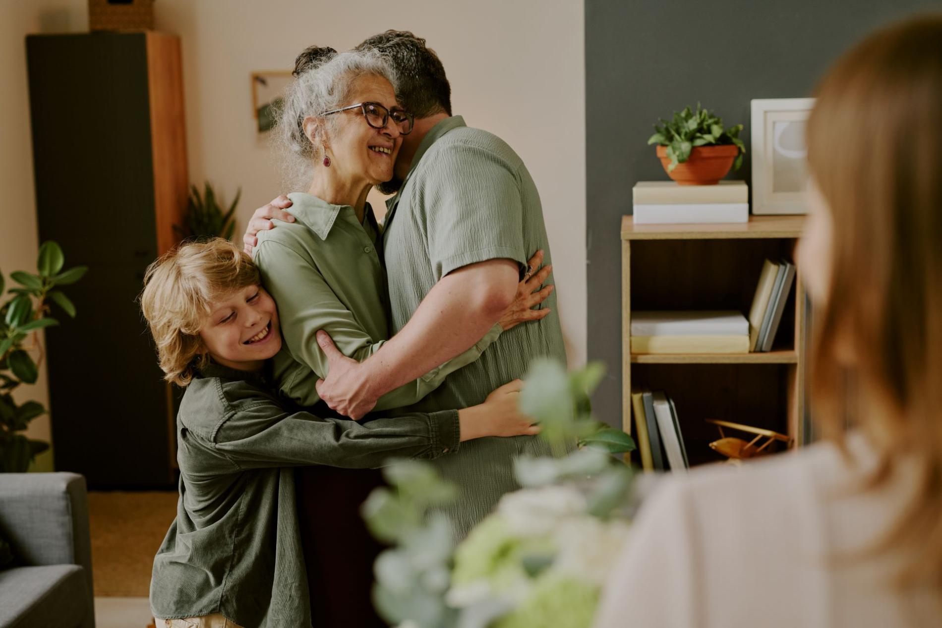 Moment d’affection intergénérationnelle : une femme âgée souriante enlace un homme adulte et un enfant dans un cadre familial chaleureux, sous le regard attendri d'une autre personne