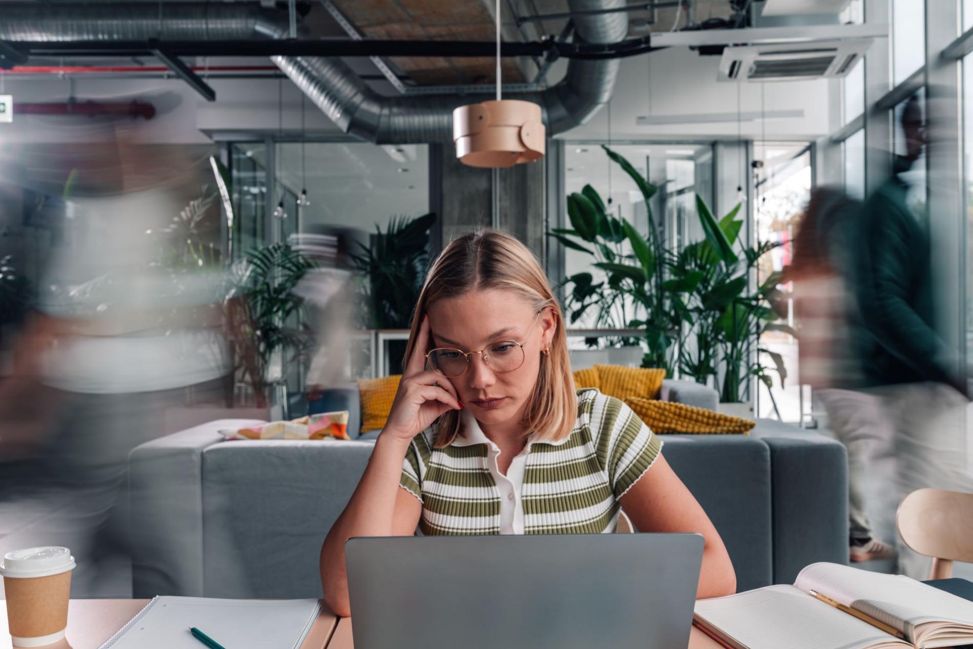Jeune femme concentrée, l’air préoccupé, travaillant sur un ordinateur portable dans un espace de coworking animé, illustrant la charge mentale ou le stress au travail chez les jeunes